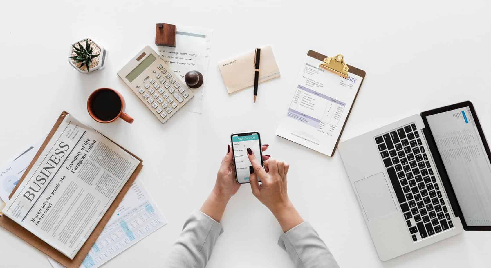 Woman holding smartphone over a white styled desk layout - passive income sources