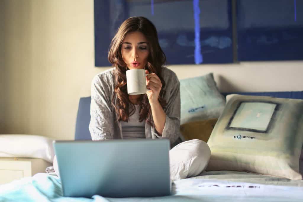 woman drinking coffee and working on laptop on bed - best transcription companies that hire beginners