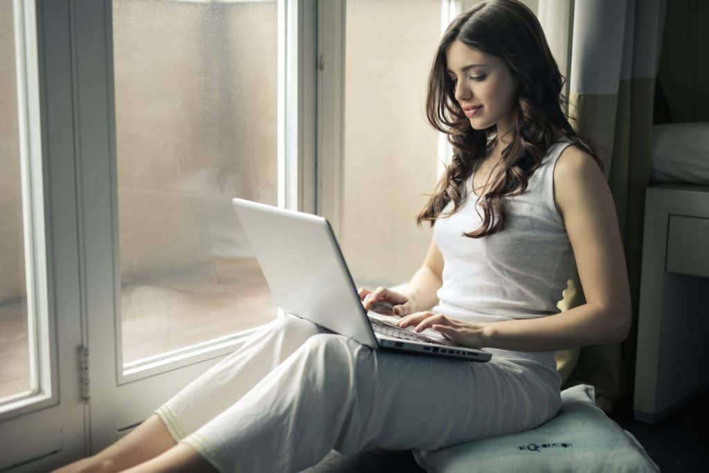 woman working on laptop by large window - best transcription companies that hire beginners