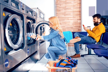 launderette near me - woman doing laundry at public laundromat and man on cellphone in background